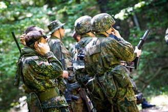Several individuals dressed in camouflage military uniforms and helmets are standing in a wooded area. Some are adjusting their equipment or communicating using radios. Sunlight filters through the leaves, creating a dappled light effect.