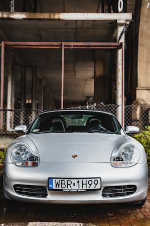 A silver sports car is parked under a concrete structure. The car has a license plate with 'PL' for Poland, indicating its location. The headlights and sleek design suggest a luxury model. Behind the car, there is a chain-link fence and an industrial background, giving the scene an urban feel.