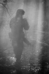 A soldier dressed in camouflage gear and wearing a helmet is cautiously moving through a dense, foggy forest. They hold a rifle ready at their side as they navigate the misty surroundings. The scene is filled with shadows and beams of light filtering through the trees, creating a mysterious and tense atmosphere.