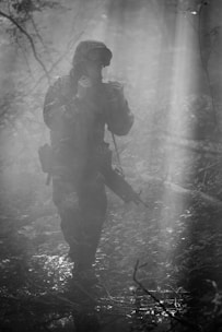 A soldier dressed in camouflage gear and wearing a helmet is cautiously moving through a dense, foggy forest. They hold a rifle ready at their side as they navigate the misty surroundings. The scene is filled with shadows and beams of light filtering through the trees, creating a mysterious and tense atmosphere.