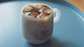 A glass mug filled with iced coffee or tea is placed on a light blue tray. The mug contains several ice cubes floating in a dark liquid topped with foam. The mug has an inscription or design on it, implying a vintage or cozy aesthetic.