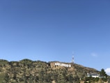 A large white sign with bold lettering is prominently displayed across a grassy and partially wooded hillside. A communications tower is visible behind the sign, and a modern-looking building sits to the right, partially obscured by trees. The sky is clear with a few wispy clouds.