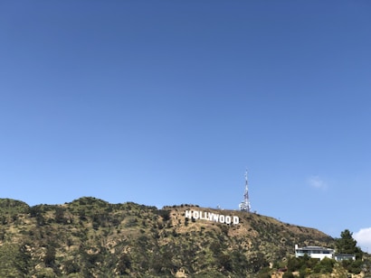 A large white sign with bold lettering is prominently displayed across a grassy and partially wooded hillside. A communications tower is visible behind the sign, and a modern-looking building sits to the right, partially obscured by trees. The sky is clear with a few wispy clouds.