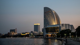 A cityscape at dusk featuring modern architecture, with a prominent sail-shaped building illuminated against the evening sky. The waterfront is lined with additional buildings, trees, and a paved walkway where a few people are visible.