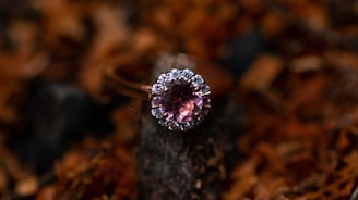 A close-up of a ring with a pink gemstone encircled by small diamonds, set against a blurred, earthy background.