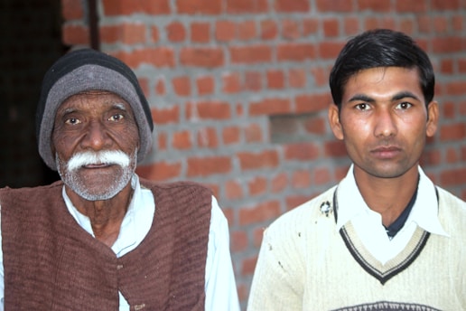 An elderly man with a white mustache and beard wears a black beanie and brown vest over a white shirt, standing beside a younger man in a light-colored sweater and white collared shirt. The background features a brick wall.