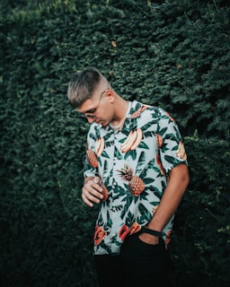 A person is standing against a dense green hedge, wearing a vibrant tropical shirt with patterns of bananas, pineapples, and leaves. The person is looking down with a relaxed posture, one hand in the pocket and the other holding a chain or accessory.