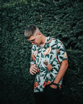 A person is standing against a dense green hedge, wearing a vibrant tropical shirt with patterns of bananas, pineapples, and leaves. The person is looking down with a relaxed posture, one hand in the pocket and the other holding a chain or accessory.