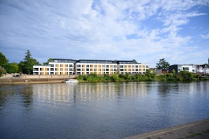 A modern apartment building with large windows sits by a calm river, surrounded by lush greenery. A small boat is moored near the riverfront, adding to the tranquil scene.
