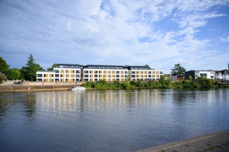 A modern apartment building with large windows sits by a calm river, surrounded by lush greenery. A small boat is moored near the riverfront, adding to the tranquil scene.