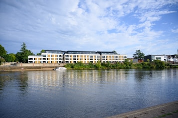 A modern apartment building with large windows sits by a calm river, surrounded by lush greenery. A small boat is moored near the riverfront, adding to the tranquil scene.