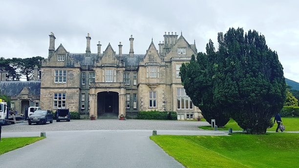 A large historic mansion with intricate stone architecture and multiple chimneys sits prominently in a well-maintained garden setting. Surrounding the mansion are neatly trimmed hedges and a large tree. Several vehicles are parked along the driveway, with a person seen walking towards the entrance.