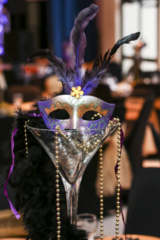A purple and silver masquerade mask with gold trim and purple feathers perched on a glass centerpiece