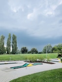 A mini-golf course surrounded by lush green grass and several tall trees under a cloudy sky. The course features geometric shapes, including yellow and green triangles. The lighting suggests a possible impending storm, with dark and thick clouds overhead.