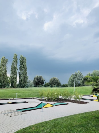 A mini-golf course surrounded by lush green grass and several tall trees under a cloudy sky. The course features geometric shapes, including yellow and green triangles. The lighting suggests a possible impending storm, with dark and thick clouds overhead.