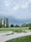 A mini-golf course surrounded by lush green grass and several tall trees under a cloudy sky. The course features geometric shapes, including yellow and green triangles. The lighting suggests a possible impending storm, with dark and thick clouds overhead.