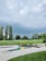 A mini-golf course surrounded by lush green grass and several tall trees under a cloudy sky. The course features geometric shapes, including yellow and green triangles. The lighting suggests a possible impending storm, with dark and thick clouds overhead.