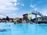 A large outdoor swimming pool with clear blue water is bustling with people enjoying a sunny day. A vibrant, winding water slide with red and yellow sections towers over the pool. People can be seen climbing the stairs to the slide and others swimming or floating in the pool. In the background, there are sun loungers and umbrellas under a partly cloudy sky, with buildings and trees providing a serene backdrop.
