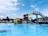 A large outdoor swimming pool with clear blue water is bustling with people enjoying a sunny day. A vibrant, winding water slide with red and yellow sections towers over the pool. People can be seen climbing the stairs to the slide and others swimming or floating in the pool. In the background, there are sun loungers and umbrellas under a partly cloudy sky, with buildings and trees providing a serene backdrop.