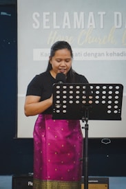 A person stands behind a microphone with a music stand in front of them, wearing a black top and a traditional patterned skirt. The background features text on a screen that indicates a welcome message related to a church setting.