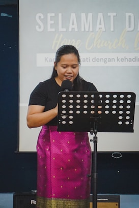 A person stands behind a microphone with a music stand in front of them, wearing a black top and a traditional patterned skirt. The background features text on a screen that indicates a welcome message related to a church setting.