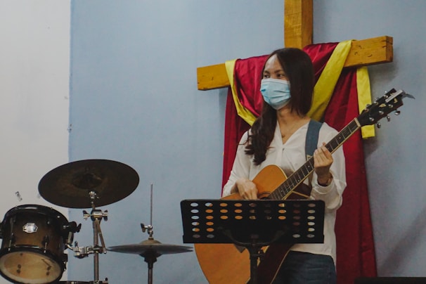 A person wearing a mask is holding a guitar in front of a microphone stand. They are standing beside a drum set and a large wooden cross draped with red and yellow cloth.