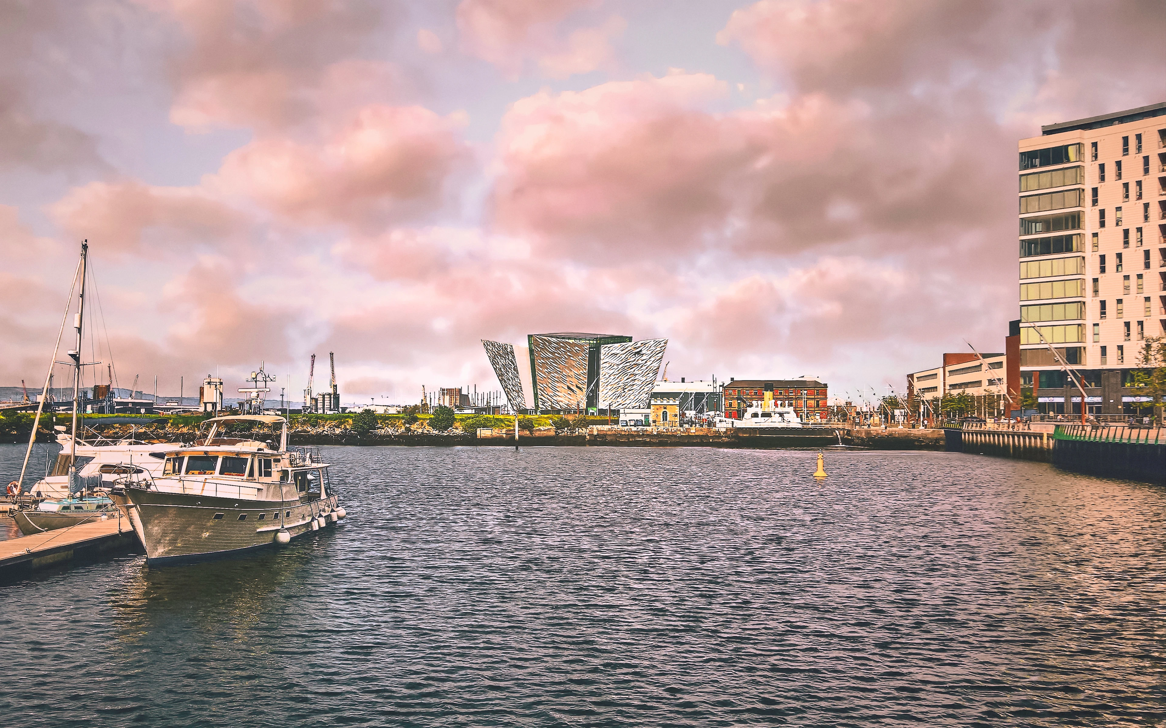 Boats moored in Titanic Quarter marina at sunset, with the Titanic Belfast building glowing under pink clouds.