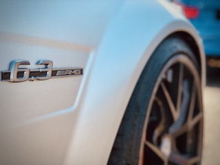 Close-up of a car's side featuring a 6.3 AMG badge with a focus on the sleek curves and design. The image includes part of the vehicle's wheel, highlighting a sporty, high-performance aesthetic.