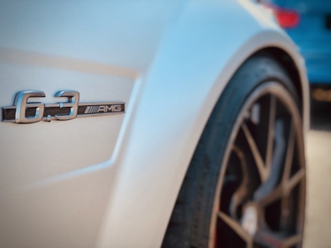 Close-up of a car's side featuring a 6.3 AMG badge with a focus on the sleek curves and design. The image includes part of the vehicle's wheel, highlighting a sporty, high-performance aesthetic.
