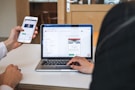 Two individuals are engaged in a collaborative setting. One person is holding a smartphone displaying a website, and the other is seated at a table with a laptop open, showing a job preference page. They seem to be reviewing or comparing information on both devices.