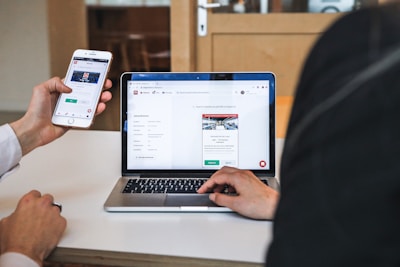 Two individuals are engaged in a collaborative setting. One person is holding a smartphone displaying a website, and the other is seated at a table with a laptop open, showing a job preference page. They seem to be reviewing or comparing information on both devices.