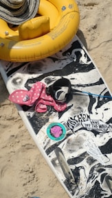 A beach scene with a surfboard that has a black and white, marbled design, adorned with stickers including a colorful one that reads 'The Beatles Surf Club'. On the sand next to the board is a pink polka dot inflatable with a floating device. A large yellow foam ring with a striped sun hat resting on top is also visible.