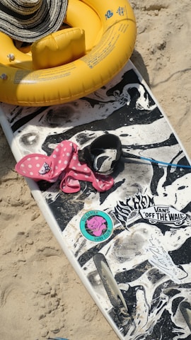 A beach scene with a surfboard that has a black and white, marbled design, adorned with stickers including a colorful one that reads 'The Beatles Surf Club'. On the sand next to the board is a pink polka dot inflatable with a floating device. A large yellow foam ring with a striped sun hat resting on top is also visible.