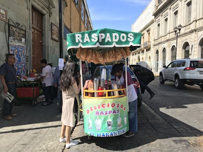 A street vendor sells raspados from a colorful cart on a busy street. Several people are gathered around the stand, one of whom is preparing the drinks. Above the cart, there are large signs advertising different flavors. The setting is a mix of traditional architecture with stone buildings in the background, and a few parked cars on the side of the street.