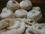 Several skeins of cream-colored yarn sit in the background, while knitting needles hold a partly-knitted fabric in the foreground. The scene suggests a cozy, crafty atmosphere.