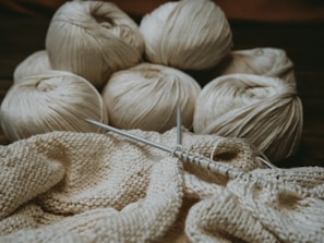 Several skeins of cream-colored yarn sit in the background, while knitting needles hold a partly-knitted fabric in the foreground. The scene suggests a cozy, crafty atmosphere.