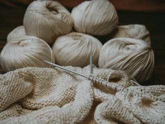 Several skeins of cream-colored yarn sit in the background, while knitting needles hold a partly-knitted fabric in the foreground. The scene suggests a cozy, crafty atmosphere.