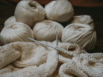 Several skeins of cream-colored yarn sit in the background, while knitting needles hold a partly-knitted fabric in the foreground. The scene suggests a cozy, crafty atmosphere.