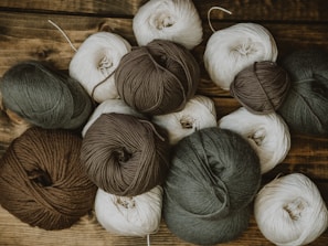 Several balls of yarn in different shades including brown, gray, and beige are arranged on a wooden surface. The yarn appears soft, with some strands loose and others tightly wound into neat balls.