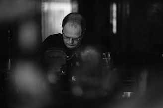 A black and white photo of a man with glasses, concentrating intently while seated indoors. The setting appears dimly lit, with blurred elements in the foreground and background, creating a sense of depth.