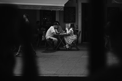 A monochrome street photograph captures a couple seated at a small outdoor café table. The woman is wearing a floral dress, and the man is dressed casually in a t-shirt and shorts. They appear to be engaged in conversation, surrounded by other patrons and passersby.