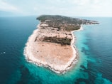 An aerial view of a rugged peninsula surrounded by clear turquoise waters. The landscape is mostly arid, with scattered vegetation and rocky terrain. A small settlement can be seen in the distance towards the narrow section of the land.