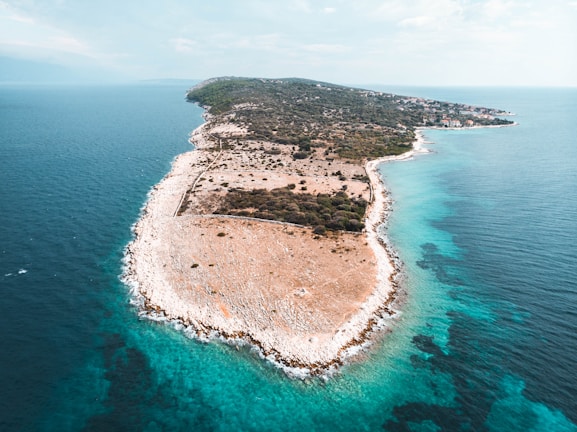 An aerial view of a rugged peninsula surrounded by clear turquoise waters. The landscape is mostly arid, with scattered vegetation and rocky terrain. A small settlement can be seen in the distance towards the narrow section of the land.