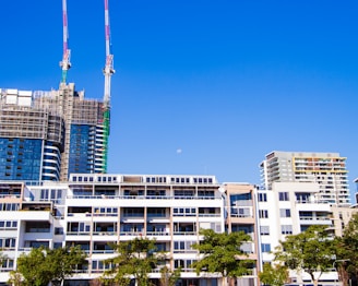 A cluster of modern high-rise buildings under construction with cranes towering above them. The architecture features a mix of glass and concrete facades. Below, there are completed residential buildings with a modern design, characterized by large windows and balconies. In the foreground, there are a few green trees, and the sky is clear with a visible moon, adding a serene contrast to the urban scene.