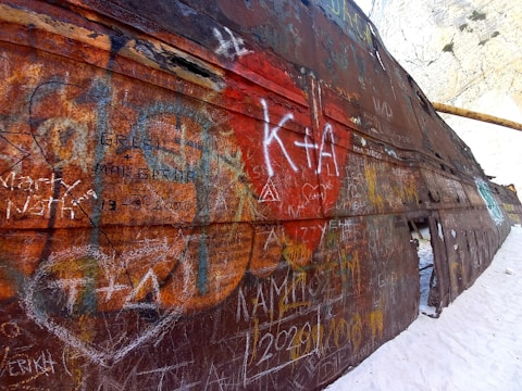 An aged, rusted shipwreck with numerous engravings and graffiti, including various names and dates, is situated on a sandy beach against a backdrop of a rocky cliff. The metal surface is heavily oxidized, displaying rich textures and layers of decay.