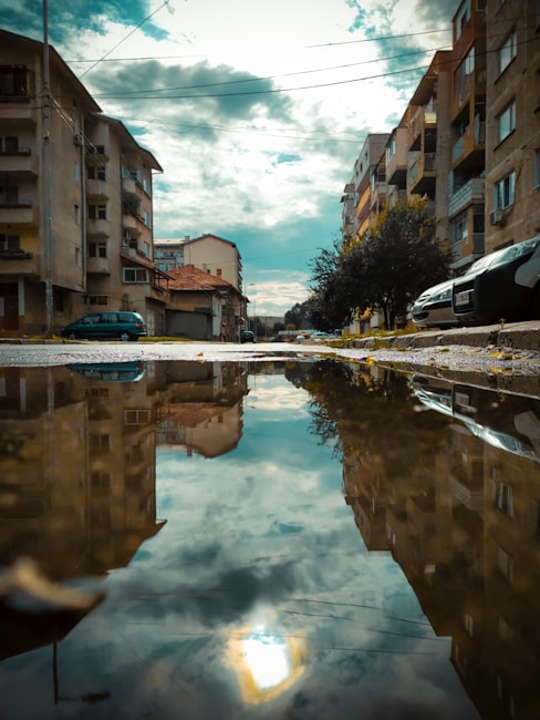 Urban scene with residential buildings lining a narrow street, featuring a large puddle in the foreground that reflects the cloudy sky. Cars are parked along the sides of the street, and power lines cross overhead.
