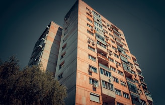 A tall, multi-story residential building with a worn, urban appearance. The facade features a mix of windows, some with balconies, and occasional satellite dishes. The perspective is angled upward with a deep blue sky in the background, and a tree partially visible at the bottom left corner.