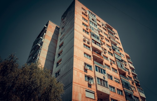 A tall, multi-story residential building with a worn, urban appearance. The facade features a mix of windows, some with balconies, and occasional satellite dishes. The perspective is angled upward with a deep blue sky in the background, and a tree partially visible at the bottom left corner.
