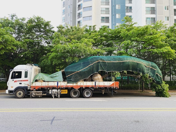 A large truck is parked on a street with a securely covered load of what appears to be a large tree, wrapped in green tarp. The background features tall residential buildings and lush green trees.