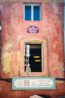A rustic storefront with an aged red facade featuring a window and a display with a small statue. Above the window, there is a sign that reads 'La Maison Rouge'. Beneath the window, a barbershop pole is visible, and a larger sign suggests various services like a barber, salon, and concept store.